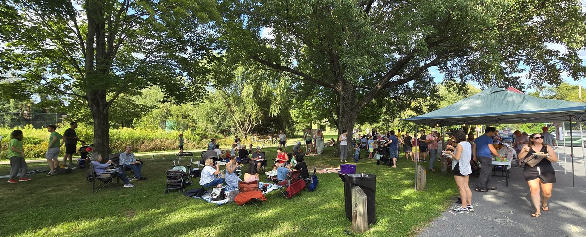 Neighbors spread across the lawn at Evans Parkway Park on a sunny afternoon, sitting in camp chairs and on blankets under big shade trees, with a line of people at the food tent along the street.