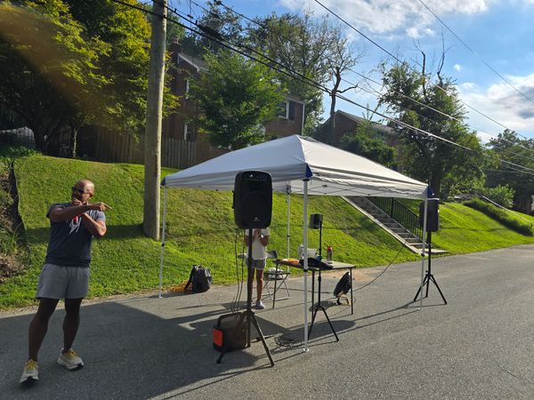 A canopy, two PA speakers on stands and a folding table set up in the middle of an empty Evans Parkway before the party, with a neighbor standing in the road.