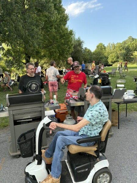 Two large grills going on the pavement with a neighbor tending them in a red shirt, another neighbor on a mobility scooter chatting alongside, and a crowd on the grass behind.
