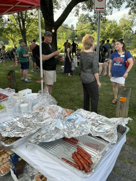 A serving table with trays of hot dogs and foil-wrapped pans, with neighbors queuing behind it on the grass.