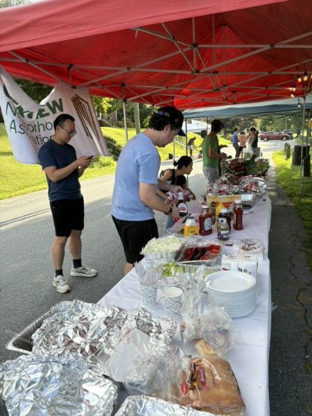 A long white table under a red canopy, covered end to end with covered dishes, condiments and stacks of plates, with neighbors serving themselves.