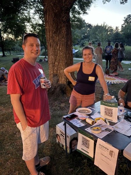Two neighbors standing at the membership table under a tree, with printed sign-up sheets and QR code cards laid out in front of them.
