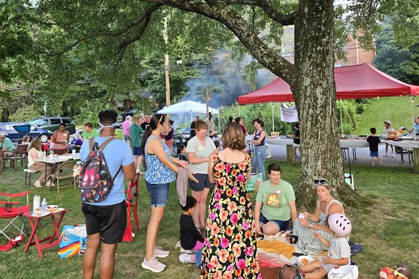Early evening under the big trees: neighbors standing and talking in groups, others sitting on blankets, with the red canopy and grill smoke behind them.