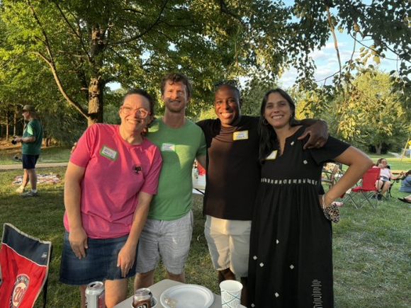 Four neighbors standing together on the grass in the evening light, name tags on, drinks in hand.