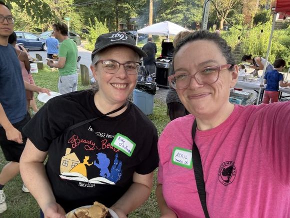 Two neighbors leaning into a photo together, wearing hand-written name tags, with the tents and the crowd behind them.