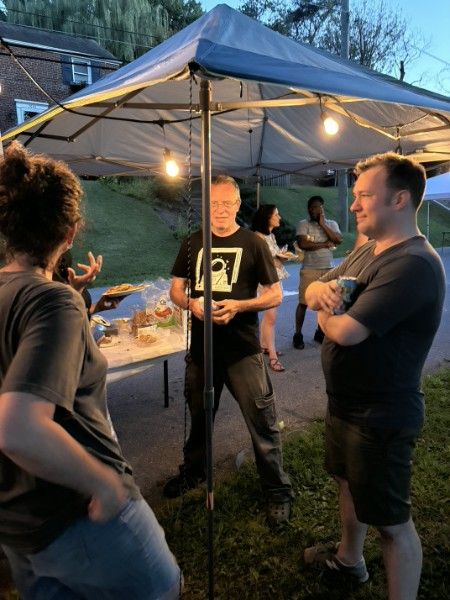 Three neighbors talking under a canopy at dusk, string lights on overhead.