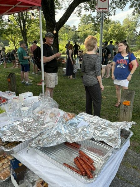 A serving table with trays of hot dogs and foil-wrapped pans, neighbors queuing behind it on the grass