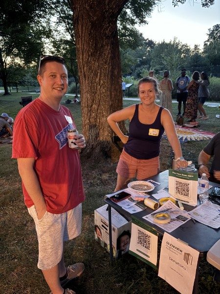 Two neighbors standing at the membership table under a tree, printed sign-up sheets and QR code cards laid out in front of them